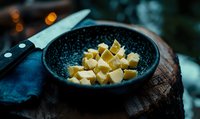 Fresh butter cubes in dark ceramic bowl on rustic wooden surface with kitchen knife and blue napkin, moody lighting and bokeh background.
