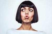 Young woman with black bob haircut making funny duck face expression against light background, wearing white top. Fashion and beauty portrait.