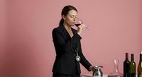 Professional female sommelier in black suit tasting red wine with eyes closed, wine bottles and glasses on table against pink background for hospitality industry.