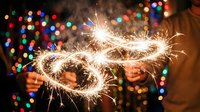Hands holding glowing sparkler forming infinity symbol against blurred colorful bokeh lights, celebrating endless possibilities and festive moments.
