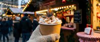 Hand in knitted glove holding steaming hot chocolate with marshmallows at festive Christmas market with bokeh lights.