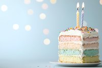 Colorful layered birthday cake slice with pastel rainbow layers, white frosting, sprinkles and two lit candles on plate against soft blue bokeh background.