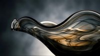 Elegant blown glass sculpture with fluid wave form, swirling amber and smoky gray tones against a dramatic dark background.