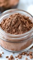 Fresh cocoa powder in glass jar with scattered powder around, macro close up shot showing rich brown texture and fine particles for baking and cooking.