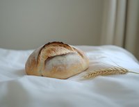 Fresh crusty artisan sourdough bread loaf with golden crust on white linen cloth, accompanied by decorative wheat stalk, natural light food photography.