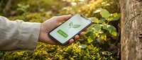 Hand holding smartphone with green leaf app screen in forest setting, eco-friendly technology concept for sustainability and nature conservation.