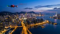 Red drone flying over illuminated urban skyline at twilight with harbor, skyscrapers, and curved waterfront highway creating stunning aerial cityscape view.