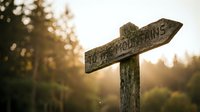Rustic wooden directional signpost pointing to mountains during golden hour sunset with warm bokeh light filtering through forest trees.