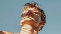 Young Caucasian man with closed eyes in dramatic sunlight creating striped shadow pattern across face against blue sky background, expressing serenity and mindfulness.