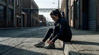 Young man with dark hair and olive complexion sitting alone on urban street curb, wearing black hoodie and sneakers, contemplative pose in industrial warehouse district setting.