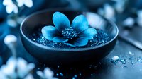 Serene blue flower floating in dark ceramic bowl with scattered petals and water drops, creating zen-like spa atmosphere against black background.
