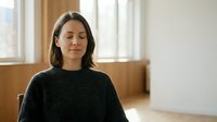 Peaceful young woman with closed eyes practicing mindfulness meditation in cozy home interior with natural sunlight streaming through windows.