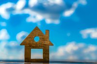 Wooden house model cutout against bright blue sky with white clouds for real estate and home ownership concepts.