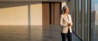 Mature woman with light skin tone, blonde hair, wearing beige blazer and white shirt, standing in modern empty office space with floor-to-ceiling windows casting shadows. Corporate professional environment.