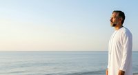 Man in white shirt standing on beach at sunset, gazing at calm ocean horizon. Suitable for wellness, lifestyle, and contemplation themes.