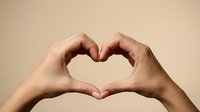 Female hands forming heart shape gesture against neutral beige background expressing love affection and romantic emotions for wellness campaigns.