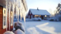 Icicles hanging from roof with snowy winter landscape and cozy cabin in background, creating serene cold weather atmosphere.