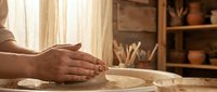 Skilled potter's hands shaping clay on spinning wheel in rustic ceramic studio workshop with natural lighting and artisan tools on wooden shelves.