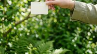 Person with light skin tone holding blank white card outdoors among lush green foliage, wearing neutral beige jacket. Suitable for business cards, eco-friendly branding, nature marketing.