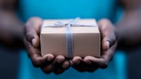 African American hands holding brown kraft paper gift box wrapped with blue twine string against dark background, offering gesture in giving position.