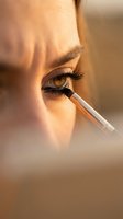 Close-up of a young Caucasian woman applying eye makeup with a brush, warm golden light highlighting her green eye and long lashes.