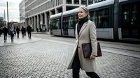 Professional businesswoman in beige coat walking through modern urban plaza with tram and office buildings in background during daytime commute.
