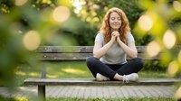 Young redhead woman sitting cross-legged on wooden park bench with hands clasped, smiling peacefully in sunny outdoor setting with bokeh background.