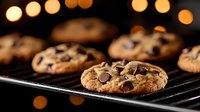 Freshly baked chocolate chip cookies cooling on black wire rack with warm bokeh lights in background, close-up view showing melted chips and golden texture.