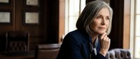 Mature woman with gray hair and fair complexion, wearing dark blazer with white collar detail, sitting in professional office setting with wooden paneling and leather furniture, contemplative expression with hand near chin, suitable for corporate, legal, business leadership, and professional services concepts.