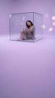 Young woman sitting inside transparent glass cube on purple background with soft bokeh lights creating dreamy surreal atmosphere for creative concepts.