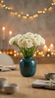White ranunculus flowers in teal ceramic vase on wooden table with warm bokeh lights creating cozy romantic atmosphere for home decor and celebration.