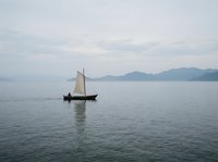 Lone sailor on a traditional wooden sailboat gliding across calm misty waters with mountain silhouettes in the background on an overcast day.