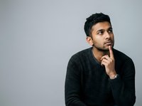 Young man with dark hair and beard, South Asian ethnicity, wearing dark charcoal sweater, touching chin thoughtfully while looking upward against neutral gray background for contemplative portraits.