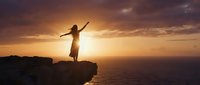 Woman silhouette with raised arms standing on cliff edge at golden sunset over ocean. Freedom, success, achievement, spiritual awakening concept.