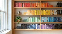 Organized bookshelf with color-coordinated books arranged in rainbow spectrum from red to purple, modern home library interior design for reading spaces.