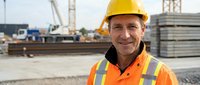 Smiling senior construction worker in safety gear at building site with cranes and materials in background, representing experienced workforce and workplace safety.