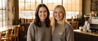 Two young women friends smiling together in cozy cafe restaurant interior with warm lighting and wooden furniture for lifestyle content.