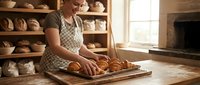 Mature woman in apron arranging fresh baked croissants on wooden board in rustic bakery kitchen with shelves of bread and natural light.