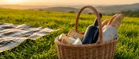 Wicker basket filled with wine bottle, bread, and cheese on green grass with picnic blanket and mountain landscape at golden hour sunset for outdoor dining and leisure activities.