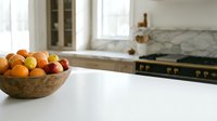 Wooden bowl filled with fresh oranges and apples on white kitchen counter with modern stove and marble backsplash in background. Fruit display.