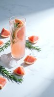 Refreshing grapefruit cocktail with rosemary garnish and ice cubes on marble surface. Fresh citrus drink for summer entertaining and healthy lifestyle.