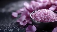 Pink Himalayan salt crystals in dark ceramic bowl with scattered rose petals on black background, spa wellness composition for aromatherapy and relaxation.