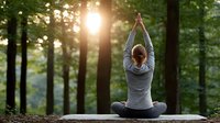 Young woman practicing yoga meditation in peaceful forest setting with warm sunlight filtering through trees creating serene outdoor wellness atmosphere.