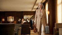 Elegant blonde businesswoman in beige coat walking through luxury hotel lobby with luggage, professional travel and hospitality concept.