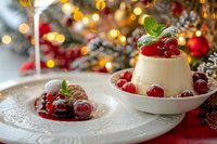 Creamy panna cotta dessert garnished with fresh cranberries, mint leaves and berry sauce on festive table with Christmas lights and decorations in background.