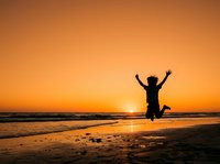 Silhouetted figure jumping on beach at sunset with arms raised in celebration, golden sky and ocean waves, freedom and joy concept for lifestyle and travel.