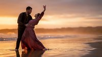 Romantic couple dancing on beach at sunset with golden sky and ocean waves, passionate embrace in flowing dress for wedding anniversary celebration.
