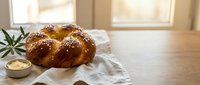 Fresh baked challah bread with sesame seeds on white cloth near sunny window, traditional Jewish braided loaf for Shabbat dinner and holiday celebrations.