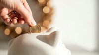 Hand inserting golden coin into white ceramic piggy bank with warm bokeh lights creating cozy savings and financial planning atmosphere.