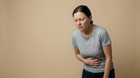 Young Caucasian woman experiencing stomach pain holding abdomen in discomfort wearing casual gray shirt against neutral beige background for health concepts.
